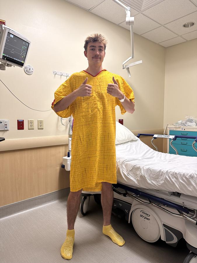 Jake in a yellow hospital gown before his second knee surgery, giving a thumbs up